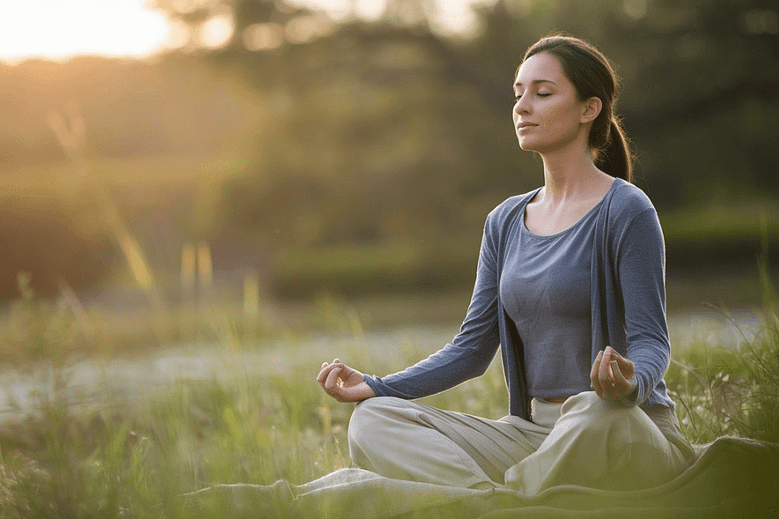 A woman practicing mindful breathing techniques outdoors, sitting cross-legged on soft grass during golden hour, surrounded by a peaceful natural environment with blurred greenery in the background.